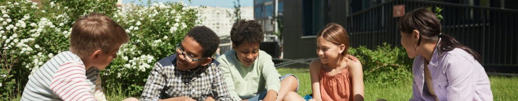School-aged children sit outside on the grass on a sunny day